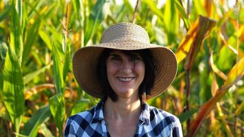 Happy smiling female farmer looks into camera standing near corn field. Portrait of adult beautiful agronomist in straw hat with maize meadow at background. Agricultural business concept. Close up photo