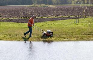 Photo of a middle-aged man mowing the grass at the pond during the beautiful spring evening.