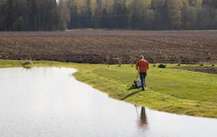 Photo of a middle-aged man mowing the grass at the pond during the beautiful spring evening.