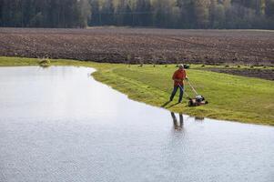 Photo of a middle-aged man mowing the grass at the pond during the beautiful spring evening.