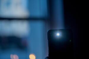 Male hand holds a smartphone with a burning flashlight with lighting window on the background photo