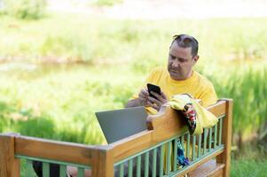 european man 50s in casual clothing sitting on bench in park and talking on cell phone while using silver laptop photo