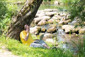 Middle aged freelancer using a laptop computer and phone while working in beautiful nature under the tree on the bank of the river photo