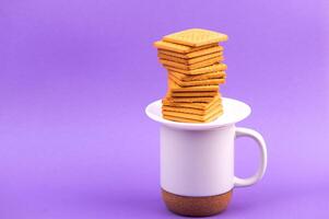 Cookies and cup of coffee. Stacks of tasty cookies on the white plate. Square biscuits on the violet background with copy space. Healthy eating photo