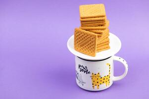 Cookies and cup of coffee. Stacks of tasty cookies on the white plate. Square biscuits on the violet background with copy space. Healthy eating photo