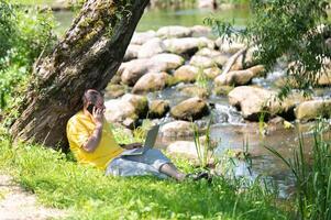 Middle aged freelancer using a laptop computer and phone while working in beautiful nature under the tree on the bank of the river photo