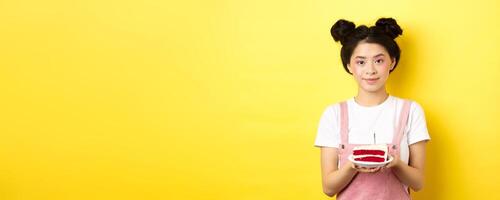 Asian birthday girl standing with cake and smiling, celebrating b-day on yellow background photo