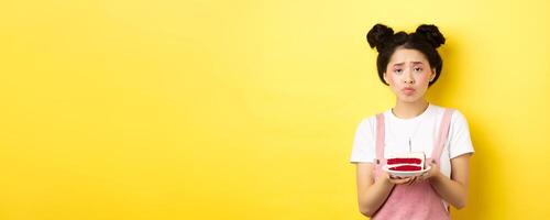 Sad and lonely birthday girl frowning upset, holding birthday cake with candle, making wish, standing on yellow background photo