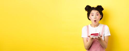 Holidays and celebration. Excited asian birthday girl blowing candle on party cake, making wish on b-day, standing against yellow background photo