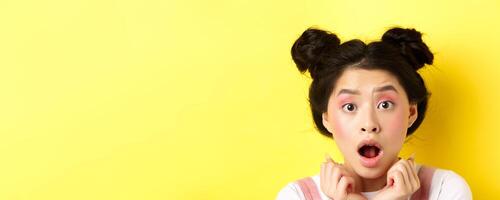 Head shot of shocked asian girl with hairbuns and glamour makeup, open mouth and looking startled at camera, standing against yellow background photo
