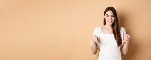 Cheerful young girl with lovely smile, pointing fingers down, making announcement or inviting click link, showing logo, beige background photo