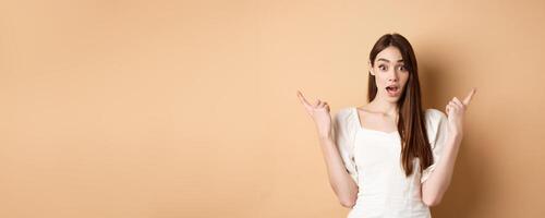 Surprised young girl pointing sideways at left and right, gasping and saying wow amazed, showing two cool choices, beige background photo