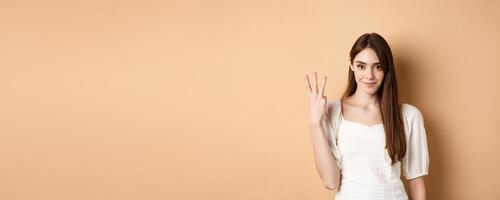 Attractive young woman show fingers number three, smiling and looking confident, standing on beige background photo