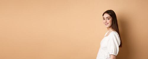 Profile of pretty european girl turn head at camera and smiling coquettish, standing in white blouse against beige background photo