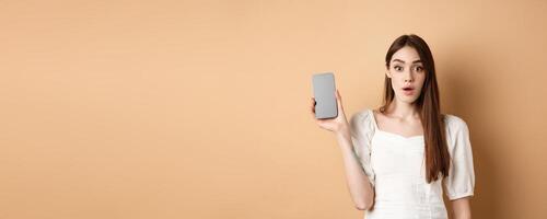 Excited pretty girl showing empty cell phone screen, look amazed at camera, standing on beige background photo