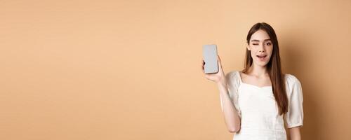 Cheerful stylish girl showing smartphone app, demonstrate empty phone screen and winking, standing on beige background photo
