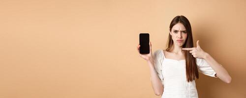 Disappointed frowning girl pointing at empty phone screen, complaining at online news, standing on beige background photo