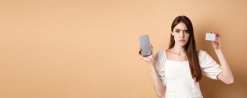 Sad and disappointed girl showing empty smartphone screen and plastic credit card, complaining and frowning, standing on beige background photo