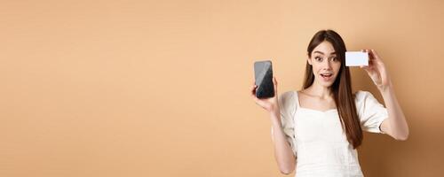 Excited girl showing plastic credit card of bank and empty mobile phone screen, demonstrate shopping app, standing on beige background photo