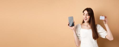 Check this out. Smiling happy young woman showing plastic credit card and empty mobile phone screen, demonstrate application, standing on beige background photo