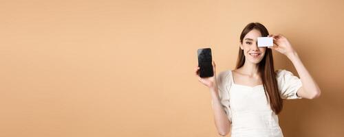 Cheerful young woman showing plastic credit card and empty mobile phone screen, smiling pleased at camera, standing on beige background photo