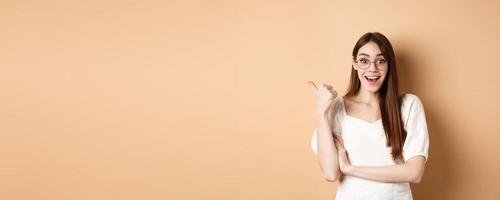 Excited girl in glasses pointing aside at logo, smiling amazed, recommending company, standing on beige background photo