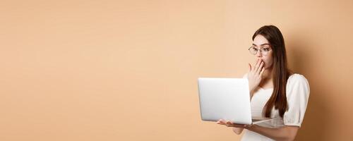 Shocked girl look at laptop screen, covering mouth with amazed face, standing in glasses on beige background photo