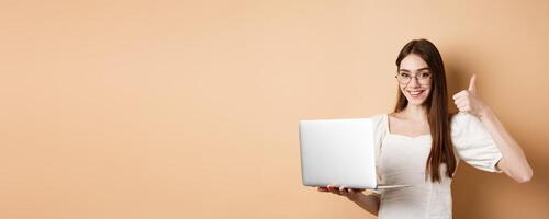 Smiling woman in glasses using laptop and showing thumb up, working on computer, standing on beige background photo