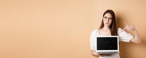 Suspicious frowning girl in glasses pointing at laptop screen, showing something strange online, standing on beige background photo