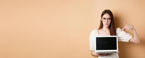 Angry frowning woman in glasses pointing at laptop screen, complaining and look disappointed, standing on beige background photo