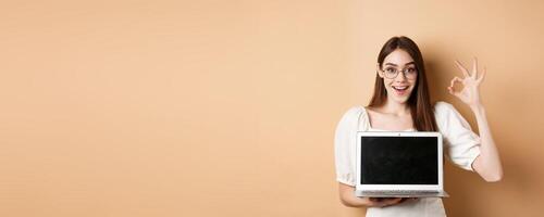 E-commerce. Excited young woman in glasses showing okay sign and laptop screen, recommending internet promo, standing on beige background photo
