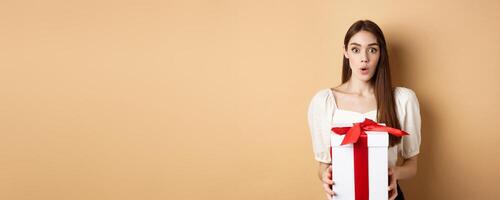 Image of surprised girl receive gift on Valentines day, looking with disbelief and holding present, standing on beige background photo