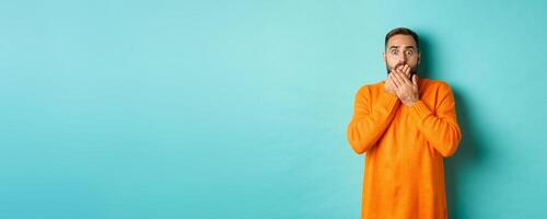 Startled man looking at horrible scene, gasping and stare scared, standing in orange winter sweater, studio background photo