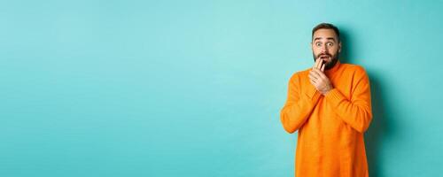 Scared and shocked guy looking at camera, standing in orange sweater against turquoise background photo
