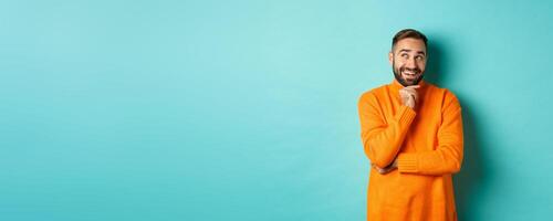 Thoughtful bearded guy making choice, shopping and looking at upper left corner, smiling satisfied, standing over turquoise background photo