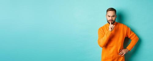 Angry bearded guy telling be quiet, hushing and frowning displeased, shut up gesture, standing over turquoise background photo