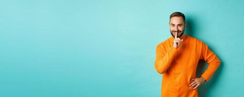 Happy man asking keep voice down, shushing and smiling, prepare surprise, hushing at camera, standing over turquoise background photo
