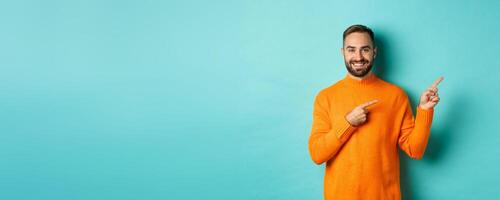 Confident happy guy in orange sweater, showing logo banner, pointing fingers right at copy space, turquoise background photo