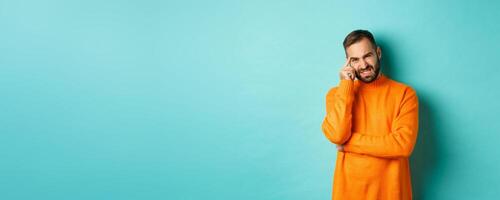 Man with beard, grimacing and cringe from something embarrassing, looking with awkward face, standing over light blue background photo