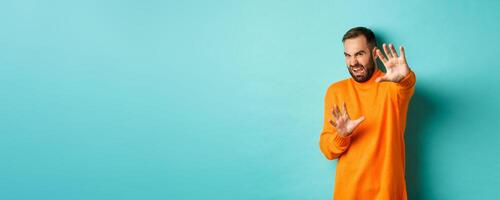 Disgusted man cringe and extending hands in protection, begging for mercy, looking at something with aversion, standing over light blue background photo