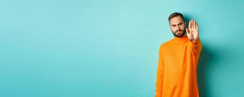 Serious frowning man showing stop gesture, telling no, prohibit and disagree, standing over light blue background photo