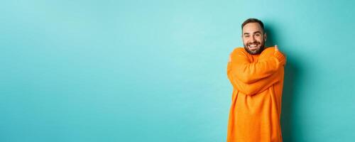 Happy man feeling comfortable, wearing warm sweater and hugging himself, smiling satisfied, standing over light blue background photo