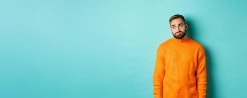 Sad and gloomy man sulking, looking bored at upper left corner, standing in orange sweater against light blue background photo
