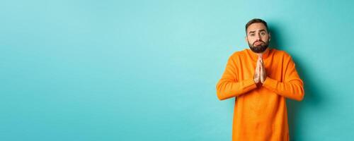 Sulking man begging for help, asking for favour, holding hands in pray and pouting, standing over turquoise background photo