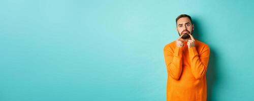 Image of gloomy bearded man, making sad face and frowning, standing upset in orange sweater against turquoise background photo