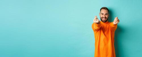 Smiling handsome man showing hearts and looking happy, lucky gesture, standing in orange sweater over light blue background photo