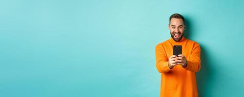 Image of man reading awesome message on mobile phone, looking at smartphone screen with amazement, standing over light blue background photo