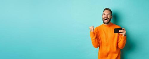 Image of man winning on mobile phone, making fist pump and rejoicing of victory, holding smartphone with satisfied face, turquoise background photo
