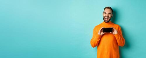 Photo of happy adult man laughing and showing funny thing on mobile phone screen, standing in orange sweater over turquoise background