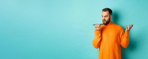 Angry man arguing on speakerphone, record voice message with mad face, standing over light blue background in orange sweater photo